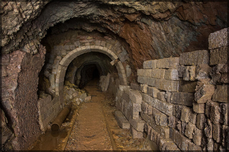 Unterirdischer Stollen mit gemauertem Gewölbebogen, freiliegendem Fels, gestapelten Steinblöcken, Gleisresten und feuchtem Boden.