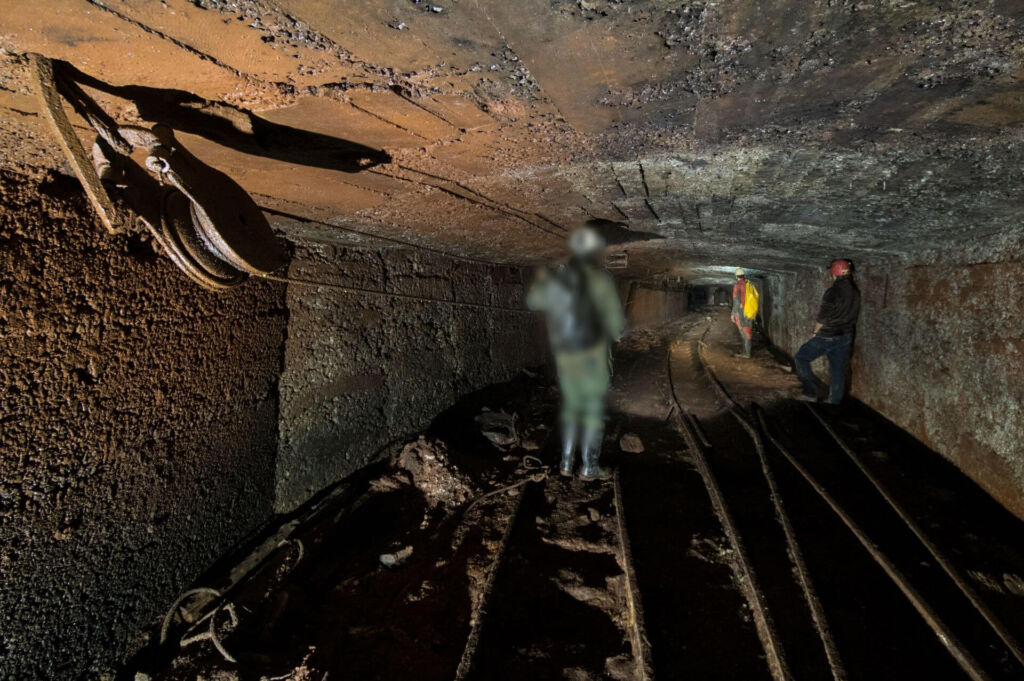Drei Personen stehen in der Ausweichstrecke und blicken in Richtung Förderschacht. Links an der Decke hängt eine große Umlenkrolle.