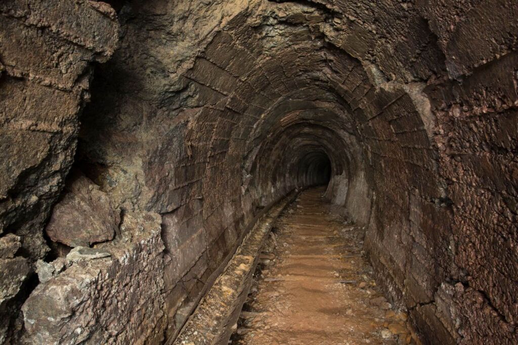 Zeigt einen Altbergbau-Stollen mit Wasserführungsrinne aus Blech. Der Stollen ist in runder Form in Beton ausgebaut.