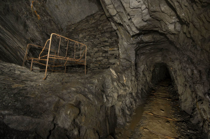Blick in einen langen Stollen und zur linken ist eine Abbaukammer zu sehen, in welcher ein Bettgestell aus Stahl vor einer Trockenmauer steht.