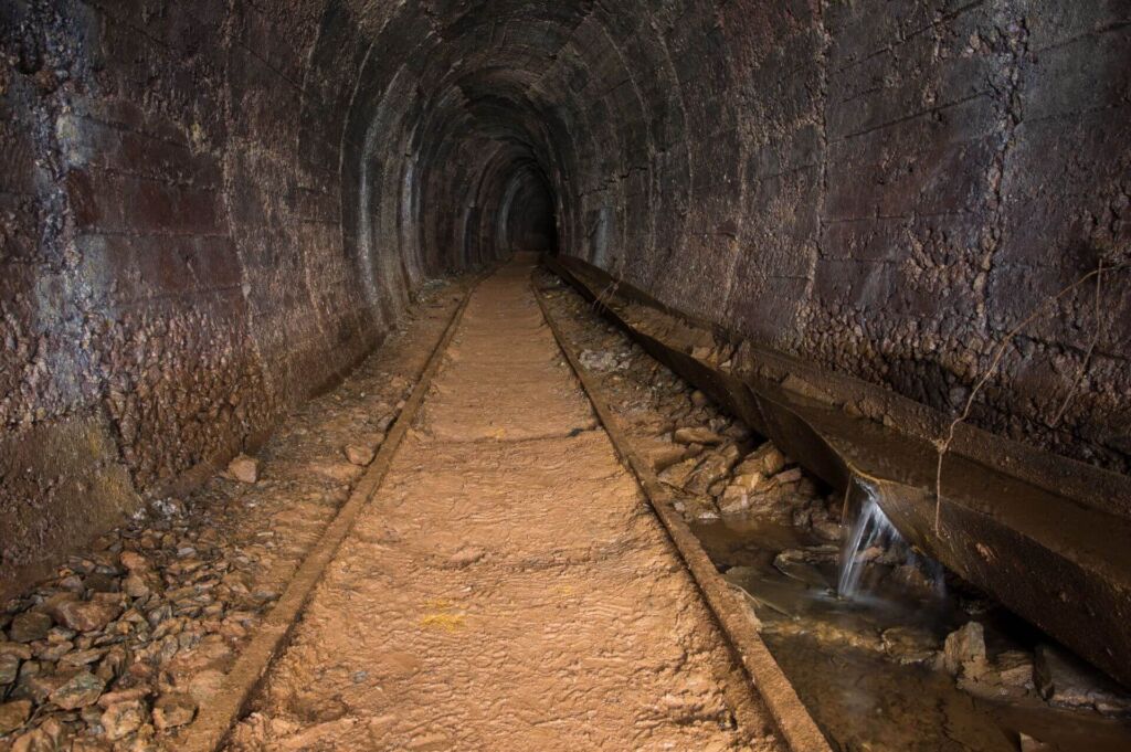 Zeigt Gleise die durch einen Altbergbau-Stollen führen. Rechts davon verläuft eine Wasserführung zum Ableiten der Grubenwässer.