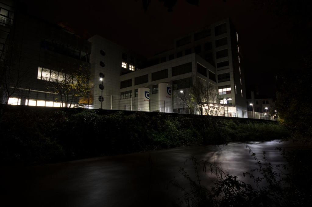 Zeigt den Fluss in der Langzeitbelichtung, im Hintergrund auf der anderen Flussseite stehen beleuchtete Industriegebäude.