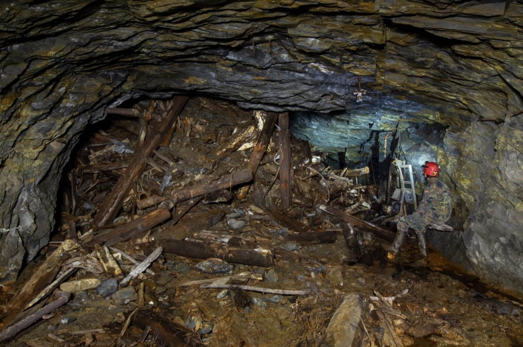 Zeigt einen riesigen Holzberg, welcher mal ein Schachtgerüst war und eingebrochen ist. Rechts auf der Bank sitzt eine Person im Stollen.