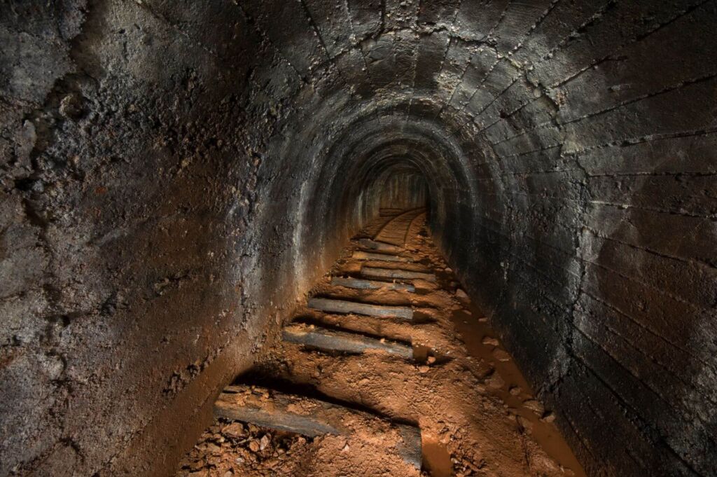 Zeigt einen Altbergbau-Stollen in runden Betonausbau. Am Boden liegen noch die Gleisschwellen und Gleise. Der Boden ist rostig rot gefärbt.
