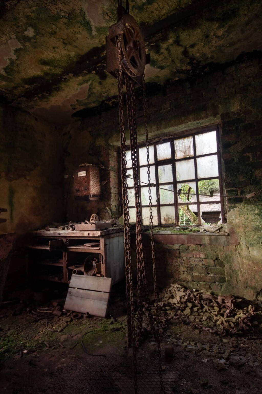 Eine kleine marode Werkstatt mit Werkbank in der linken Ecke. Die Wände sind moosig grün und von der Decke hängt ein Flaschenzug.