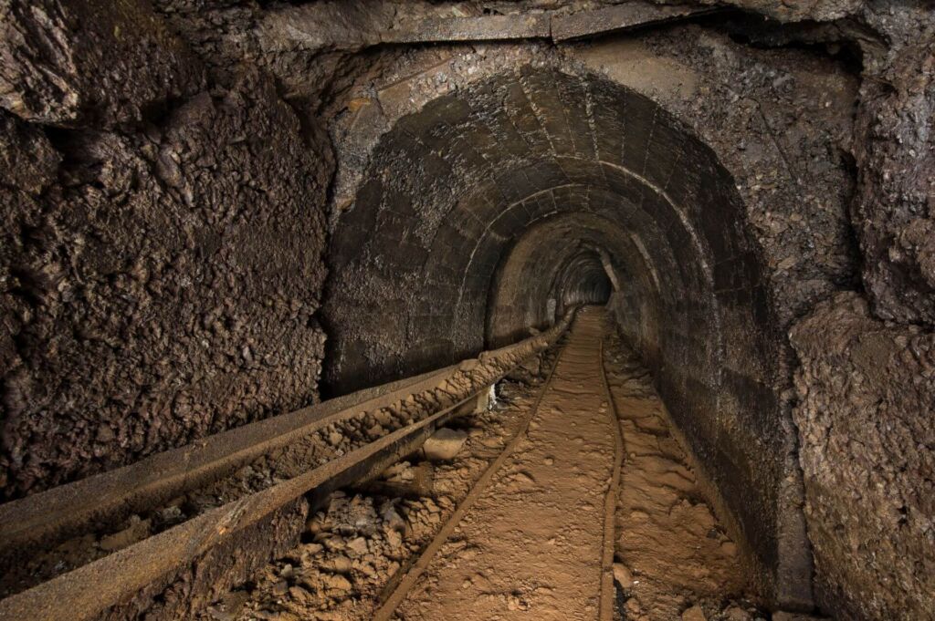 Zeiht einen Altbergbau-Stollen in Betonausbau, mit Gleisen am Boden und einer Wasserführung am linken Stoß. Die Erde leuchtet braun durch den hohen Eisengehalt.