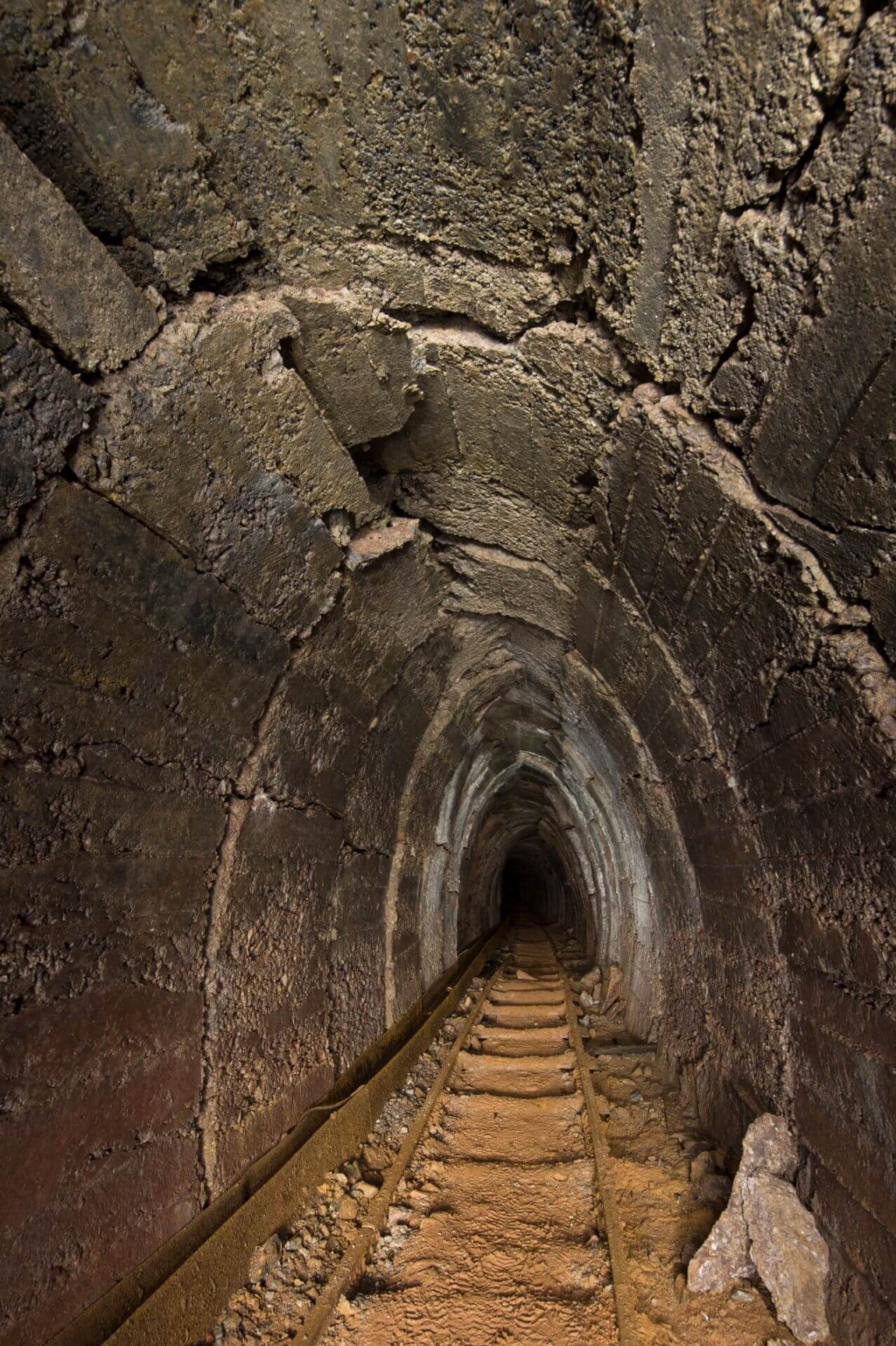 Zeiht einen Altbergbau-Stollen in Betonausbau, mit Gleisen am Boden und einer Wasserführung am linken Stoß. Der Beton an der Firste ist vom Bergdruck geborsten.