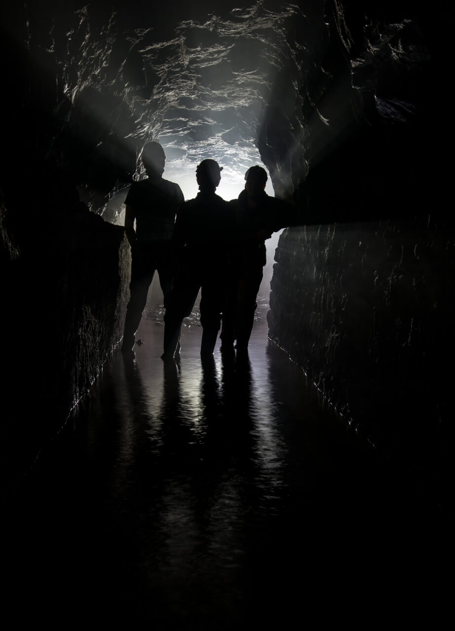 019-tortenstollen Zeigt drei Personen im Gegenlicht, wie sie im Stollen stehen. Das Wasser steht ihnen bis zum Knöchel.