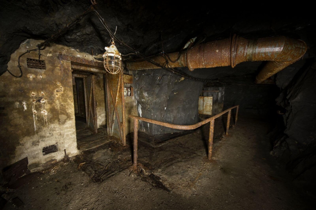 Zeigt einen dunklen, schroffen Stollen. An der Decke verläuft ein dickes rostiges Lüftungsrohr zum Sprengstoffbunker.