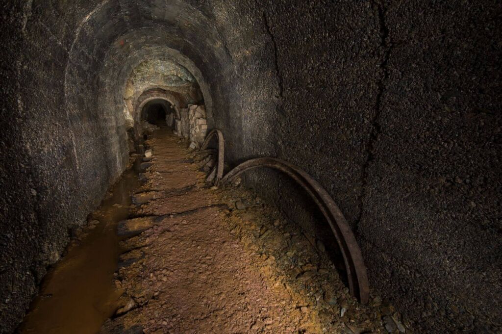 Zeigt einen Altbergbau-Stollen, welcher in Beton ausgebaut wurde. Links fließt das Grubenwasser und rechts an der Wand stehen Stahlteile.