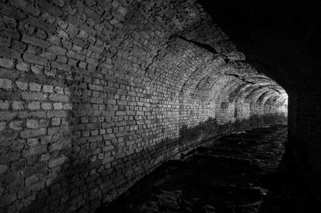Zeigt eine in Ziegel ausgebaute Strecke im Altbergbau. Aus der Ferne strahlt Licht und am Boden liegen die Gleisschwellen.