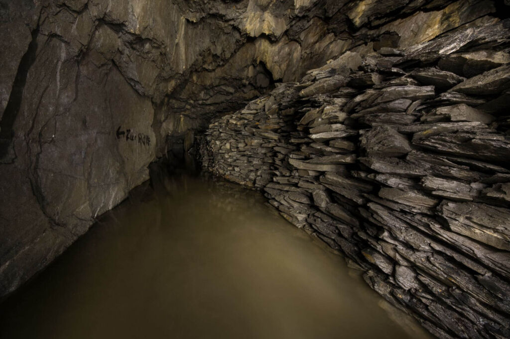 Zeigt eine lange Versatzmauer aus Schieferplatten. Der Stollen steht Kniehoch mit Wasser voll und am linken Stoß steht mit schwarzer Farbe geschrieben "Zur Hölle"