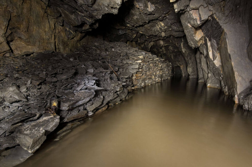 Zeigt einen Schieferabbau, welcher kniehoch mit Wasser voll steht. Links eine Trockenmauer, welche dahinter mit Versatzmaterial gefüllt.