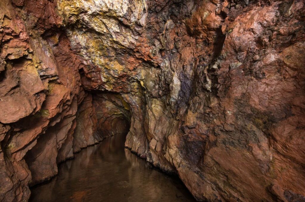 Zeigt einen Streckenabschnitt, in dem 20 cm Wasser steht. Die Wände sind schroff und rot gefärbt. An der Firste ist das Erzband erkennbar.