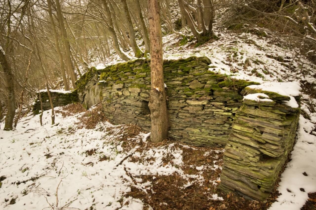 Zeigt eine Grundmauer aus Schiefersteinen am Hang, welche zu einem Grubengebäude gehörte.
