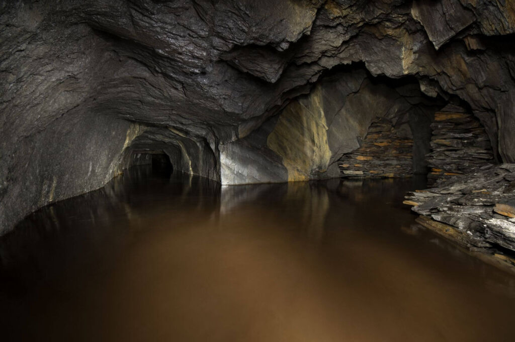 Zeigt einen Stollen, der bis zur Hälfte unter Wasser steht. Rechts im Bild ein Schieferabbau mit Trockenmauerung.
