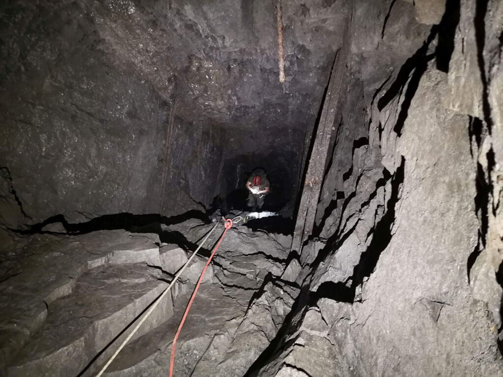 Zeigt einen tiefen Schacht im Schiefergestein. Etwa 10 Meter tiefer hängt eine Person im Seil und steigt auf.