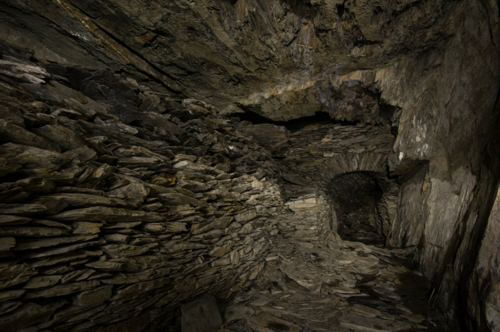 Zeigt eine große Trockenmauer aus Schiefersteinen, im Anschluss einen Durchgang aus Schiefer, der im weiteren Verlauf verbrochen ist.