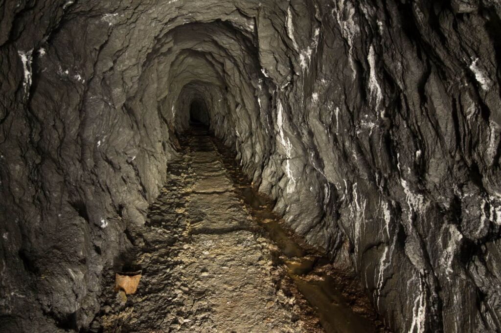 Zeigt einen langen grauen Schieferstollen, in dem rechts am Boden das Wasser fließt. Vereinzelt findet sich Sinter am rechten Stoß.