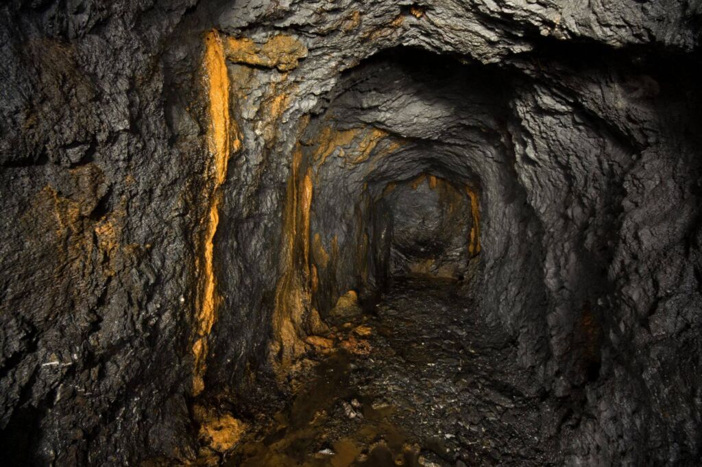 Zeigt einen Schieferstollen mit orangefarbigen Versinterungen am linken Stoß.