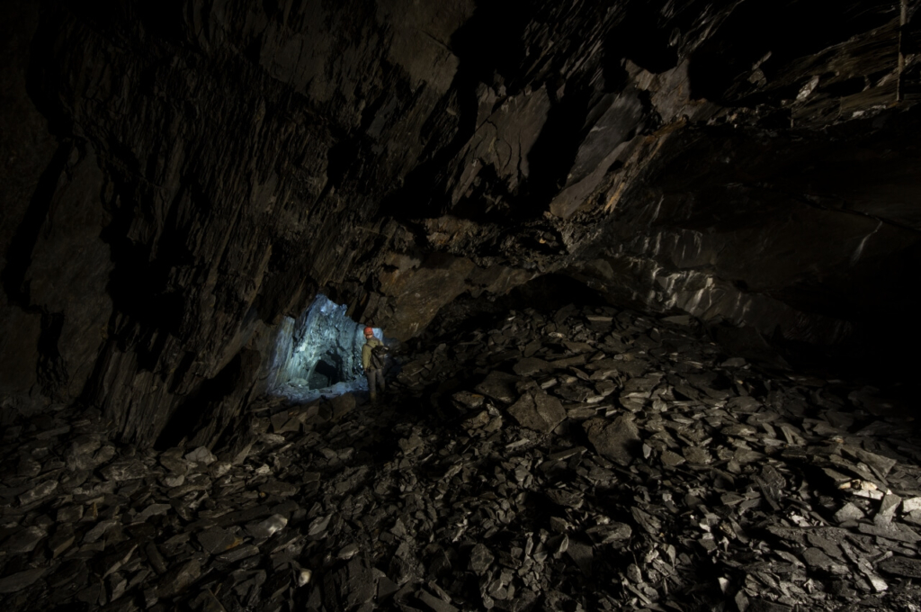 Zeigt einen großen Hohlraum, in dem Schiefer abgebaut wurde. Weiter entfernt steht eine Person und leuchtet in den anschließenden Stollen.
