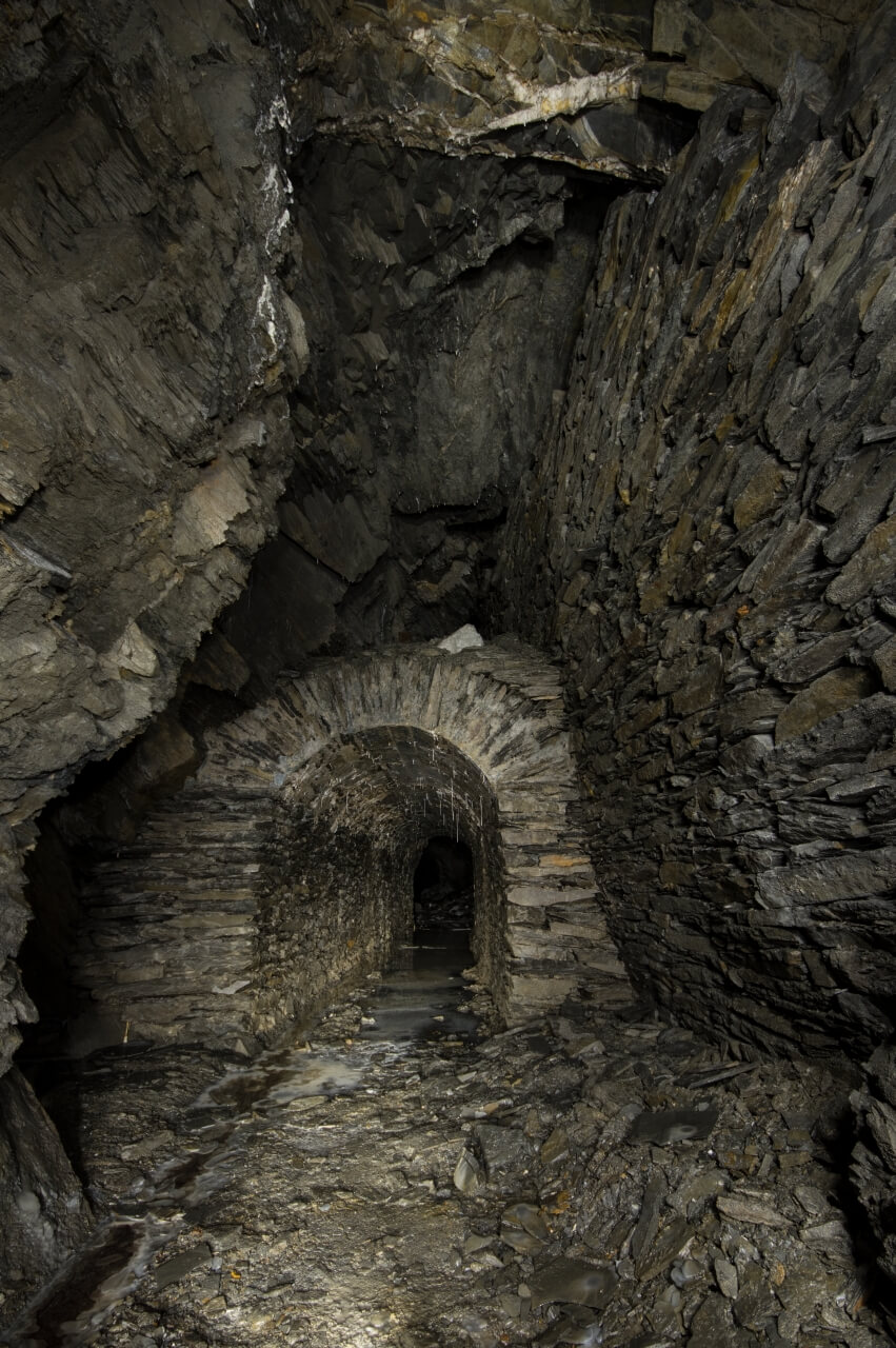 Zeigt eine Bogenmauerung aus Schieferstein, der Gang führt durch den Abbau. Der Boden ist mit weißem Sinter bedeckt.