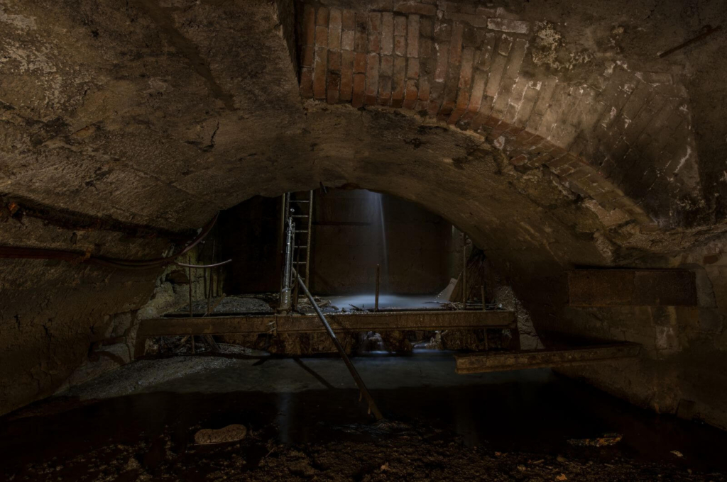 Zeigt ein Gewölbe mit einer Stahlleiter. Von oben kommt Licht und viel Wasser läuft herunter.
