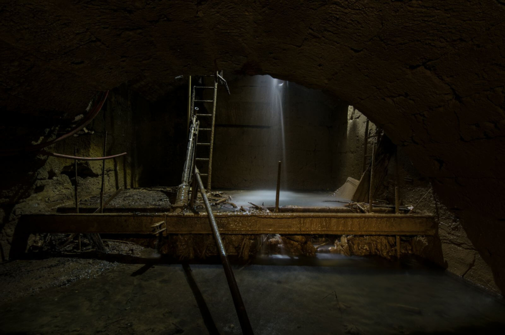 Zeigt ein Gewölbe mit einer Stahlleiter. Von oben kommt Licht und viel Wasser läuft herunter.