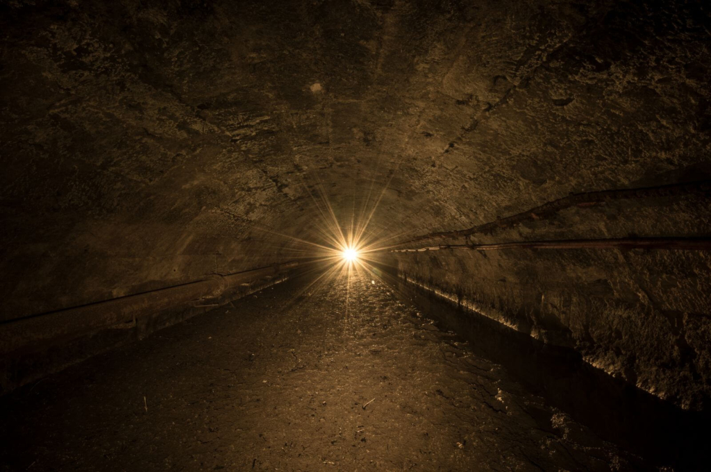 Zeigt eine unterirdische Flussumleitung. Die Decke ist rund und an der linken Seite verläuft eine Wasserleitung. Das Flussbett ist schlammig und nur rechts läuft noch etwas Wasser.