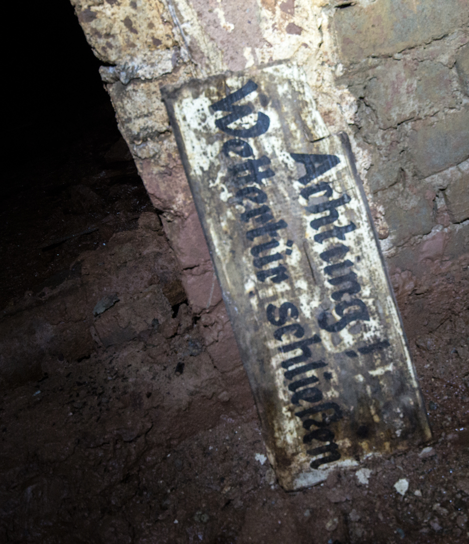 008_wettertuer_schließen_detail Zeigt ein Holzschild, das an einer Ziegelmauer lehnt, mit der Aufschrift "Achtung! Wettertür schließen".