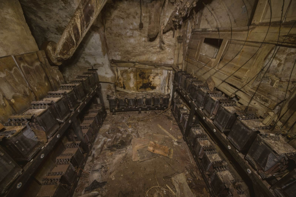 Zeigt den gesamten Raum mit seinen Bleiakkus. Links, rechts und geradeaus stehen morsche Regale mit den alten Bleiakkus.