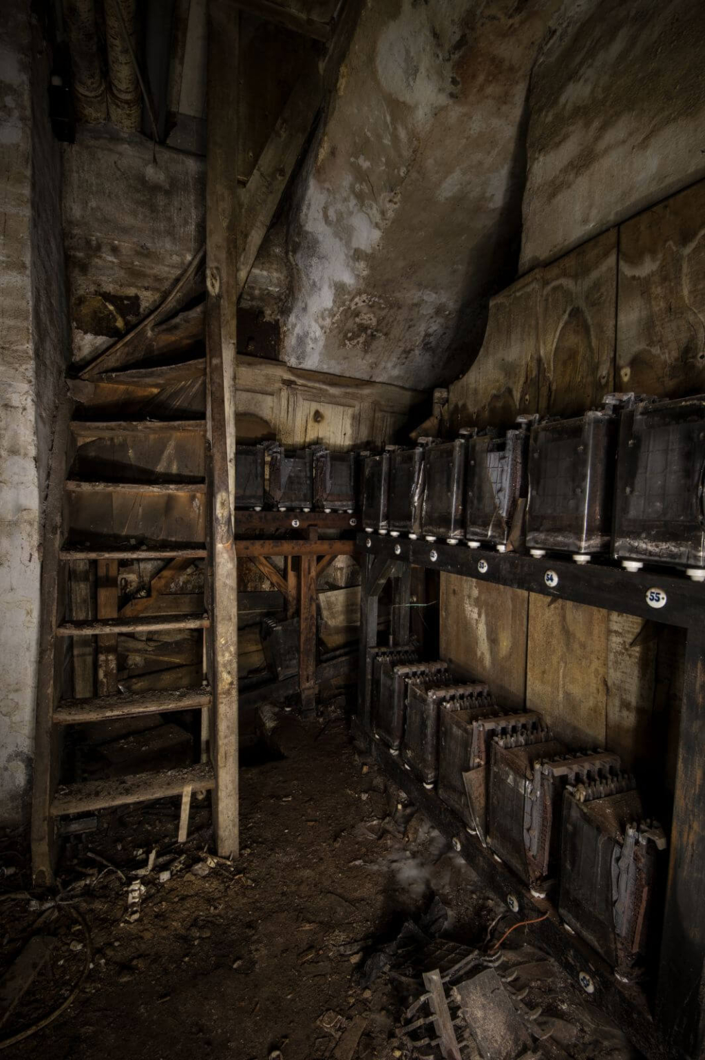 Zeigt einen verlassenen Raum, voll mit alten Akkumulatoren. auf morschen Regalen. Links führt eine Treppe nach oben.