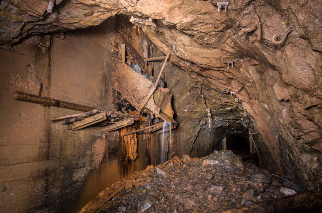 013_wasserrolle Zeigt eine eiserne Rollenschnauze im Stollen, vor der ein Berg Geröll liegt. Aus der Rollenschnauze läuft Wasser.
