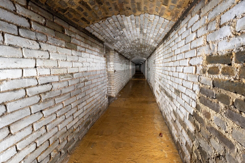 Langer Flur mit links angrenzenden Schutzraumtüren und nass, schlammigen Boden.