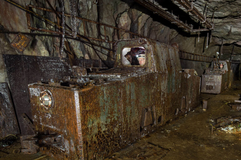 Rostige Grubenlokomotive in einem unterirdischen Stollen, umgeben von Felswänden, Schienen, Fördertechnik und hängenden Leitungen, mit einer Person im Führerstand.