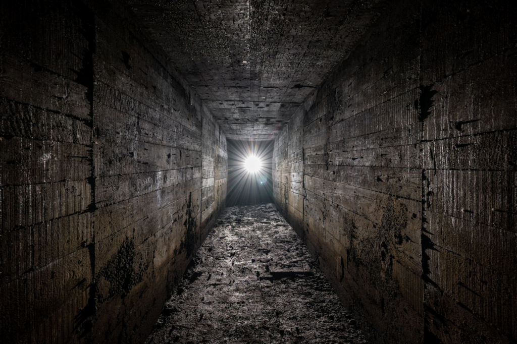 Rechteckiger Zugangsstollen aus Beton im Gegenlicht. Der Boden ist matschig und an der Decke hängen Tropfen.