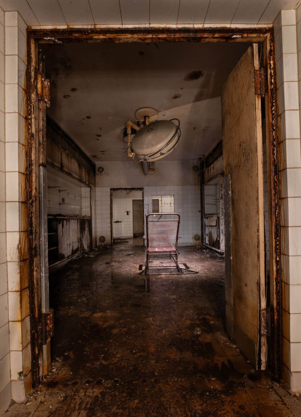 Blick durch die Tür in einen OP-Raum mit Liege in der Mitte des Raums und leuchtender OP-Lampe darüber.