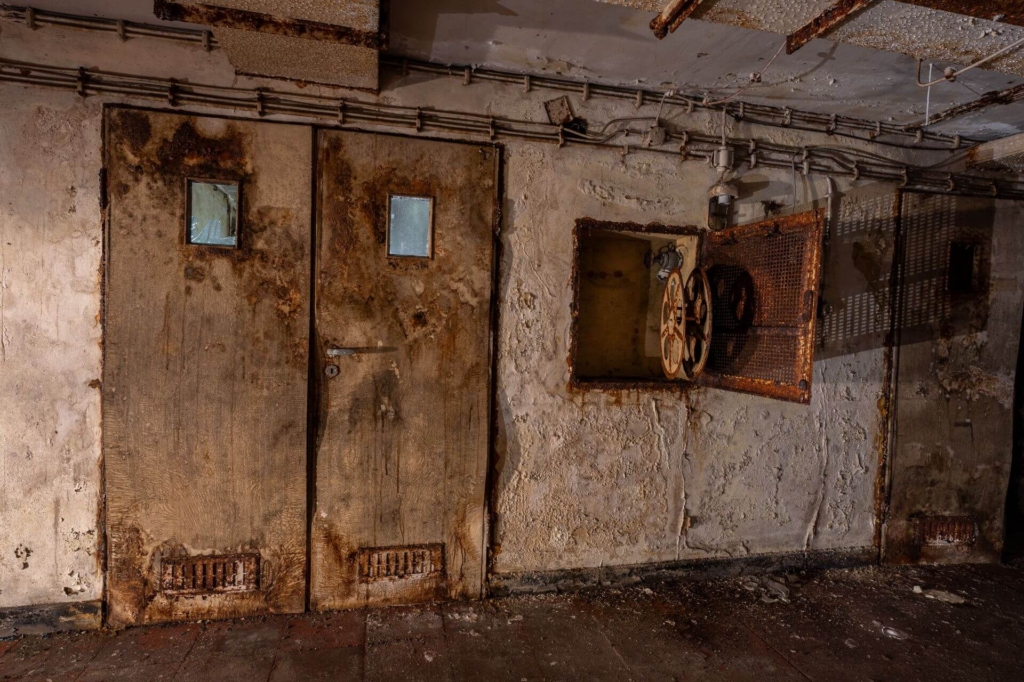 Modrige Flügeltür zum Patientenzimmer mit kleinen Fenstern, rechts daneben Nische für Feuerwehrschlauch.