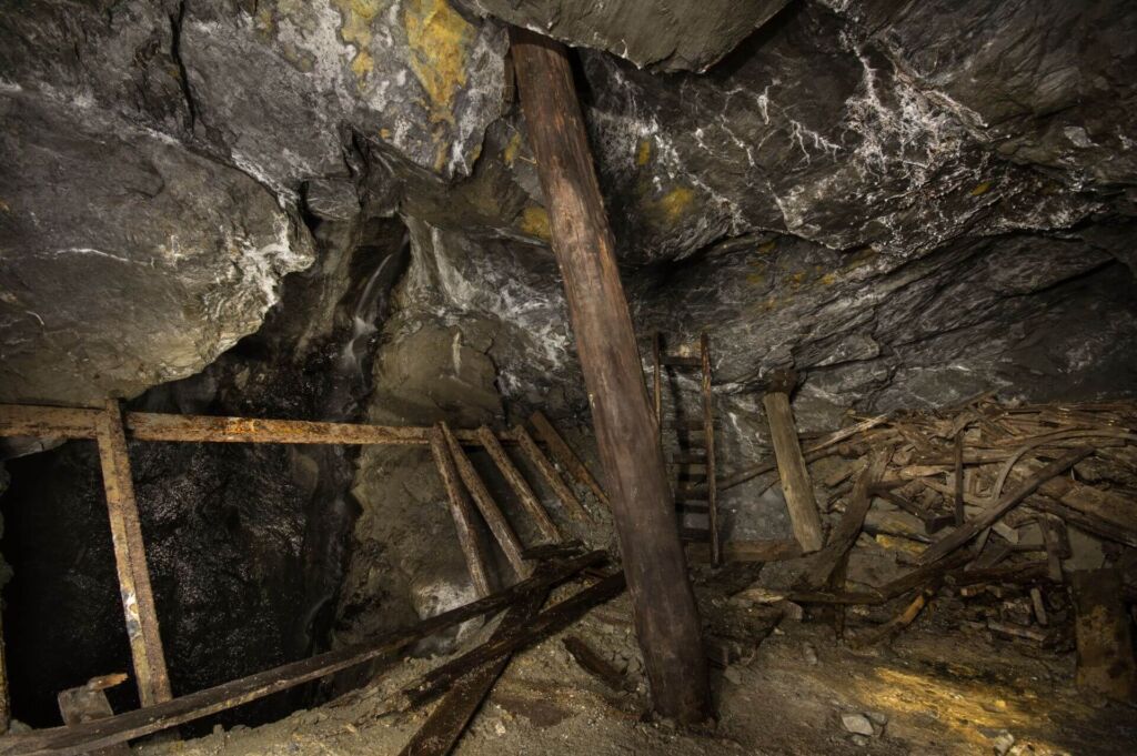 010-altbergbau-stempel-am-schacht Links ein Rollentrichter und in der Mitte steht ein Stempel, der die Firste stützt. Rechts liegen viele Bretter und Hölzer auf einem Haufen.