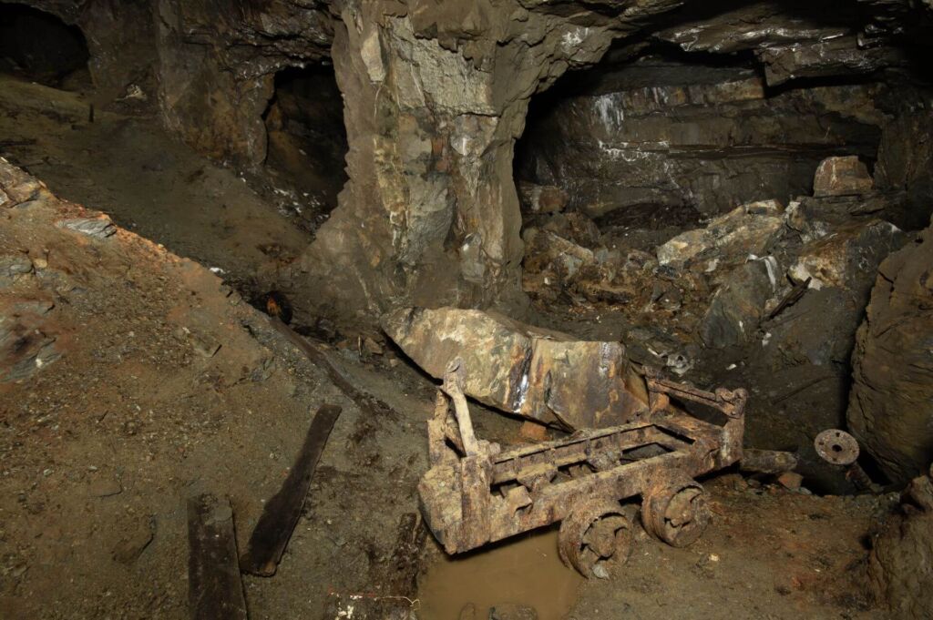 015-altbergbau-foerderwagen-rahmen Zeigt ein rostiges Lorenchassi, das im Pfeilerkammerbau steht. Rechts im Bild liegen große Felsen, die aus der Firste gefallen sind.