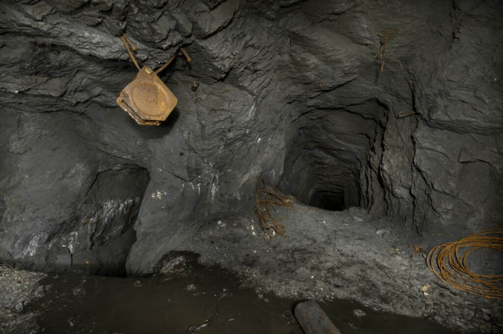016-altbergbau-eisenerzgrube-umlenkrolle-schrappergasse Zeigt zwei Stollen, die schräg nach unten verlaufen. An der Decke hängt eine rostige Umlenkrolle und am Boden liegt ein aufgerolltes Stahlseil.
