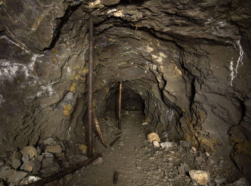 019-altbergbau-eisenerzgrube-stollen-stempel Zeigt einen Stollen, der auf eine Leiter zuführt. Es liegt marodes Holz auf dem Boden.