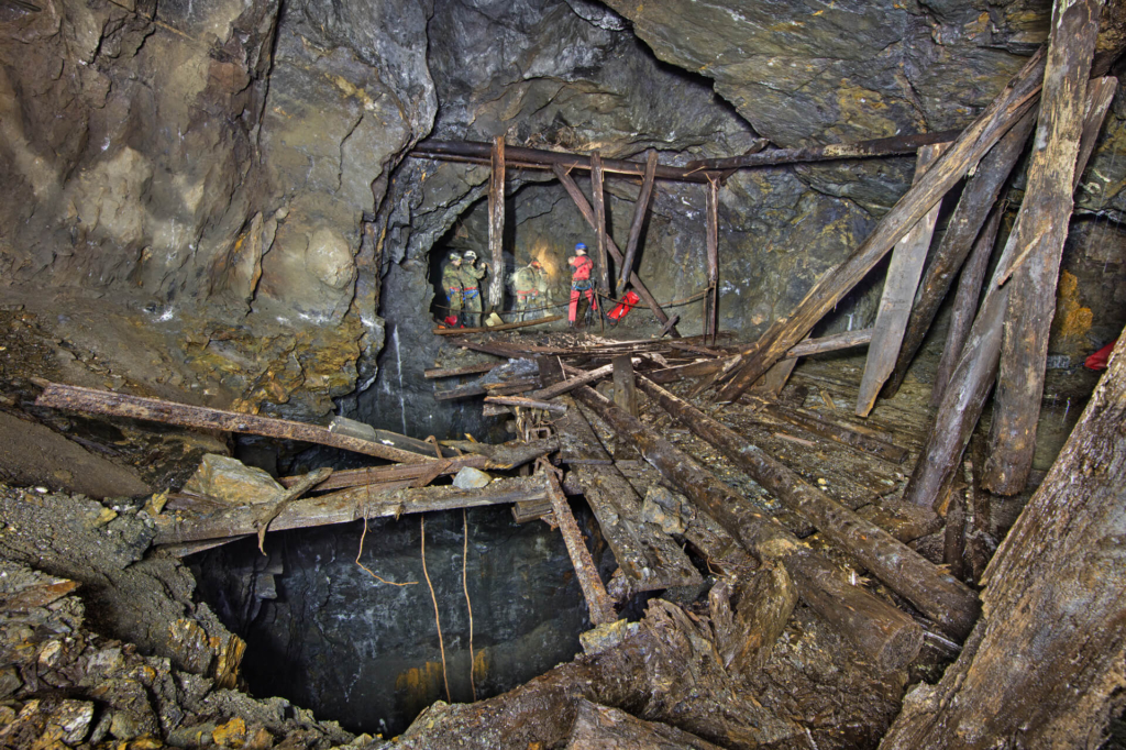 020-altbergbau-eisenerzgrube-schmiedefeld-abseilstelle Vorne im Bild ein tiefer Rollentrichter, dahinter stehen vier Personen, die ein Seil zum Abseilen einbauen.