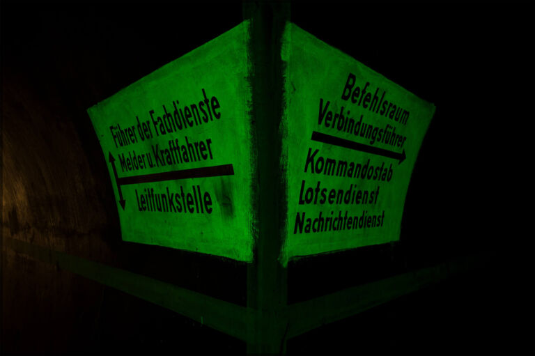 Leuchtend grüne Wegweiser im Luftschutzstollen unter dem Stuttgarter Killesberg mit Beschriftungen zu Befehlsraum, Kommandostab, Nachrichten- und Lotsendienst sowie Leitfunkstelle, aufgenommen im Tunnelambiente.