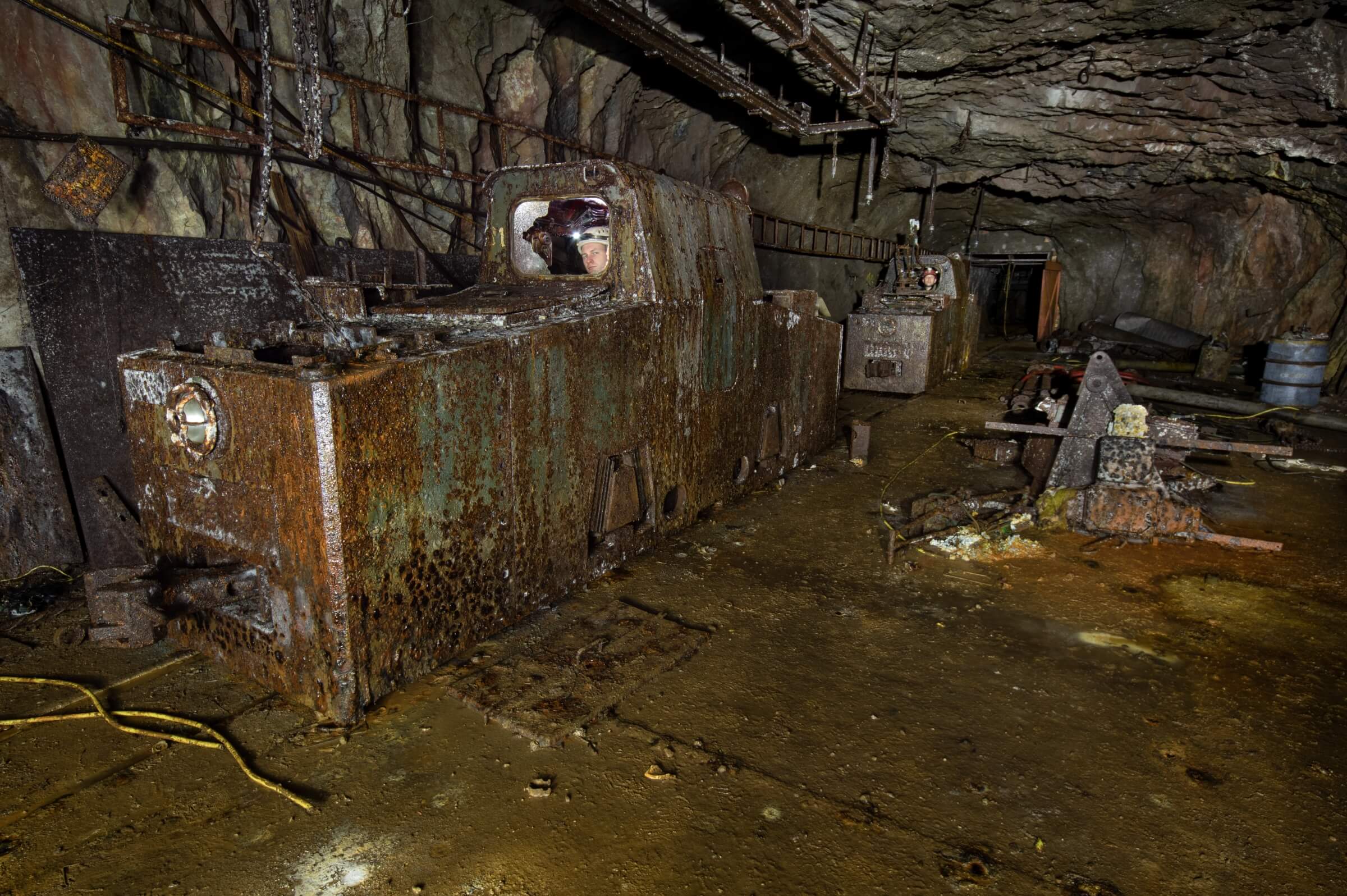 Eine verrostete Fahrdrahtlokomotive von der Firma AEG steht in einem verlassenen Bergwerkstollen, umgeben von alten Ausrüstungen.